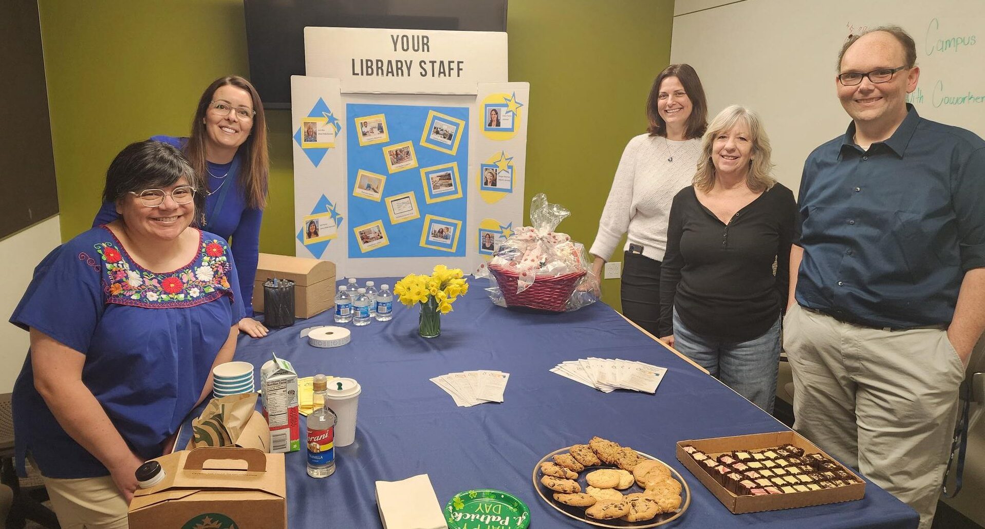 Library staff posing with President and VP in front of library informational poster and a table of snacks